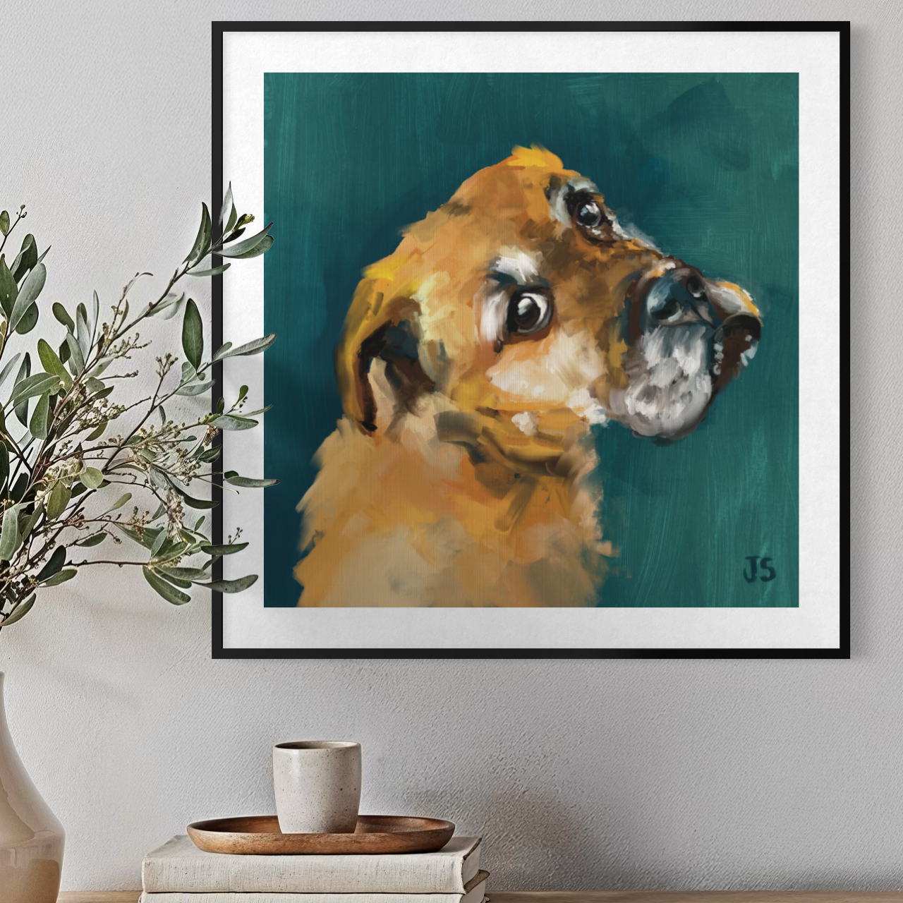 Framed wall art featuring a painted portrait of a golden dog on a teal background, displayed in a black frame on a light gray wall above a table with books, a cup, and decorative branches.