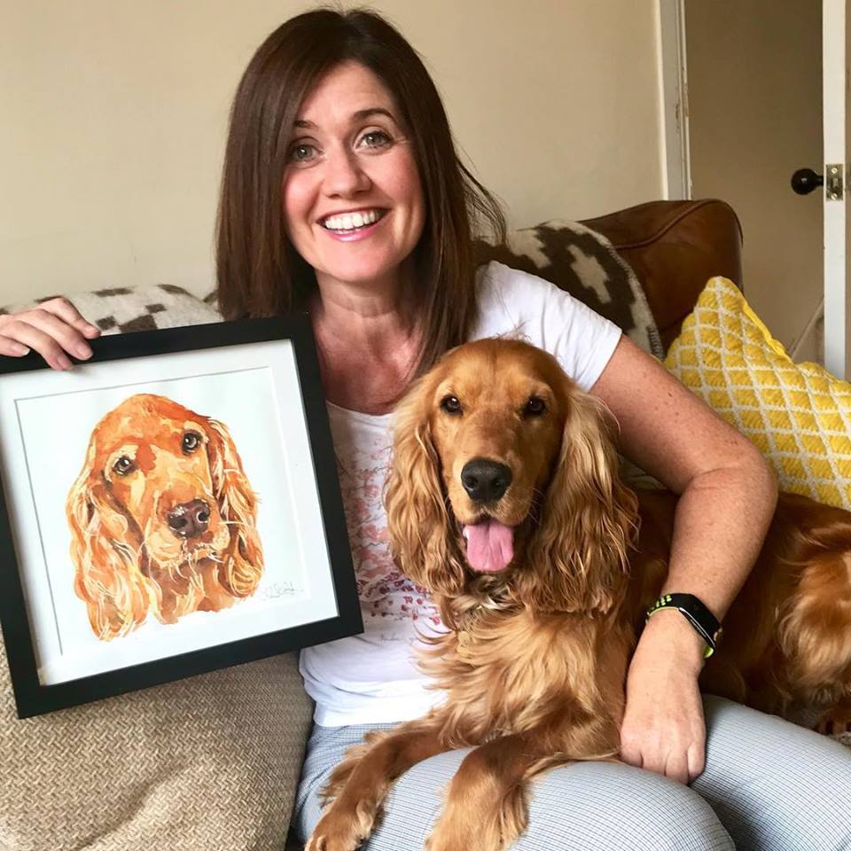 Photograph of a smiling customer holding a water colour portrait of their golden cocker spaniel dog who is sat on their knee