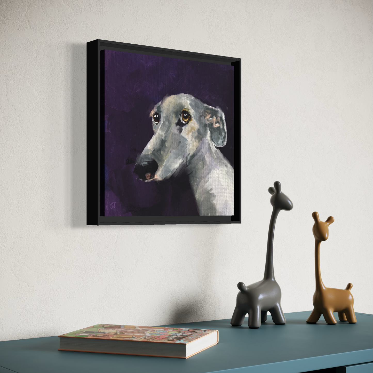 A framed portrait painting of a greyhound dog on a dark purple background, displayed on a wall above a blue table with decorative giraffe figurines and a book.