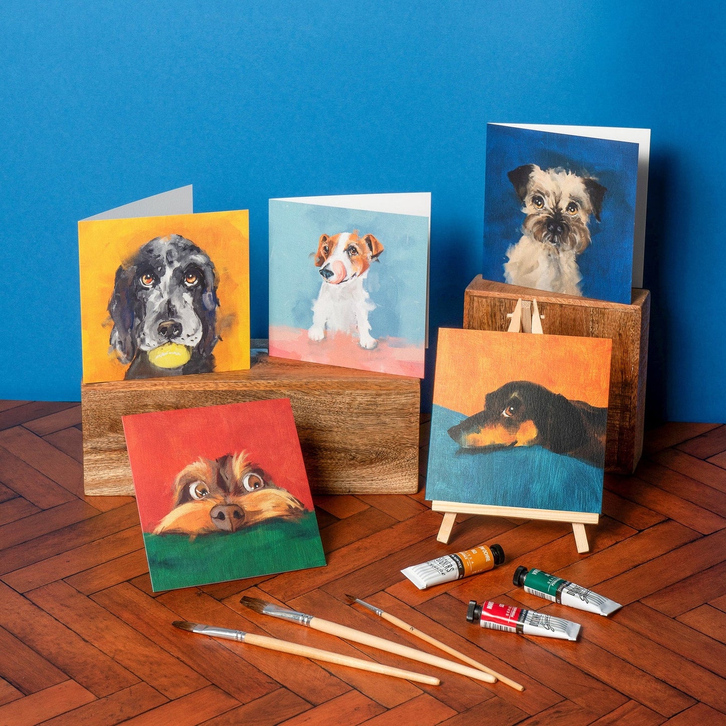 Collection of dog-themed greeting cards and paintings on a wooden surface with a blue wall background.